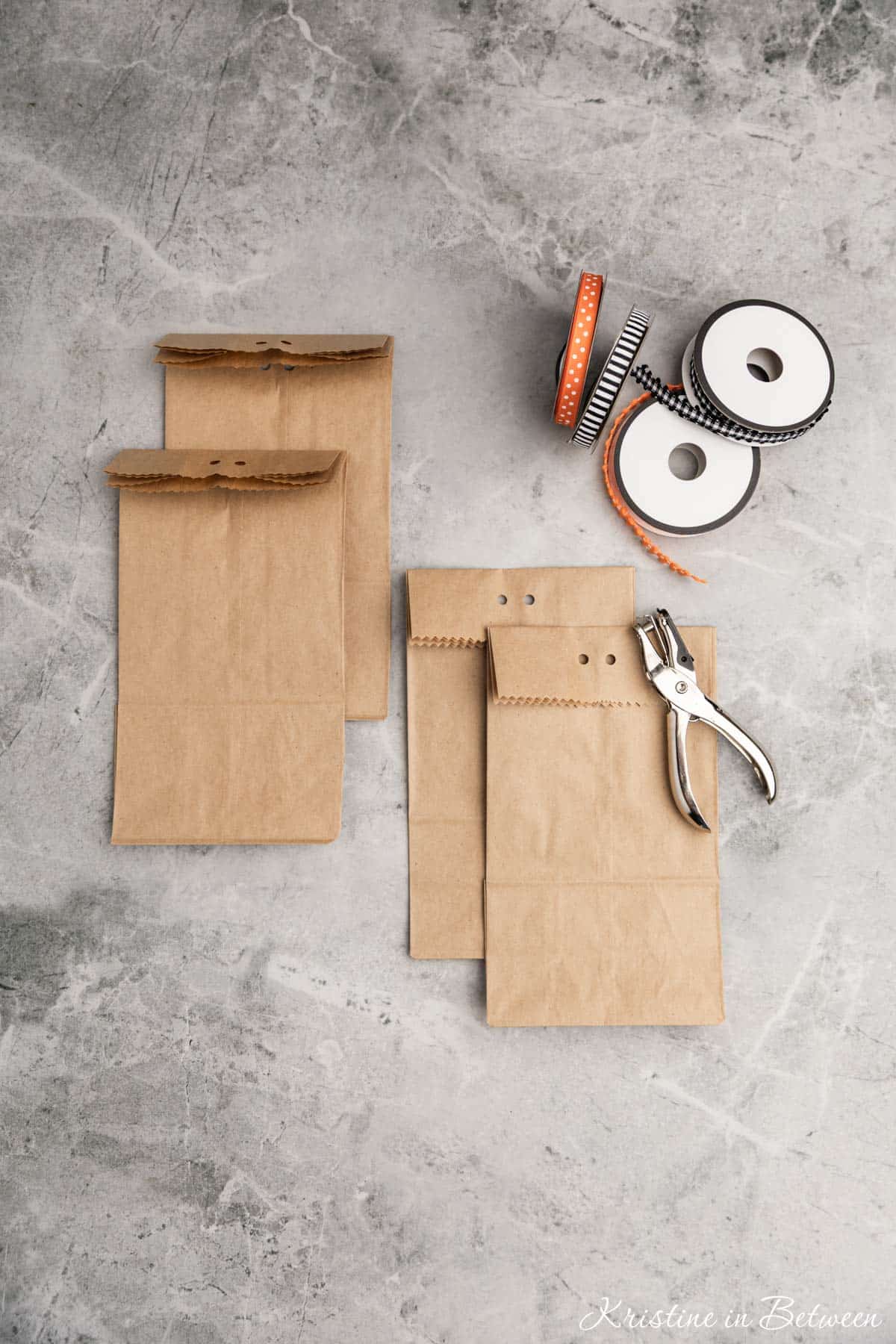 Brown paper bags folded down with two holes punched in the top laying with a hole punch and ribbons.