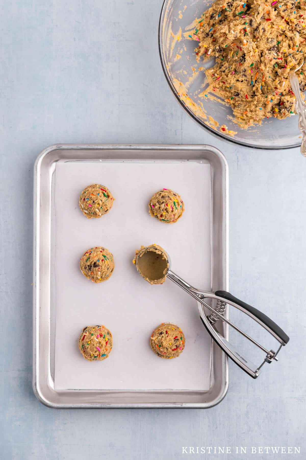 Scooped cookies on a lined baking sheet sitting with a cookie scoop.