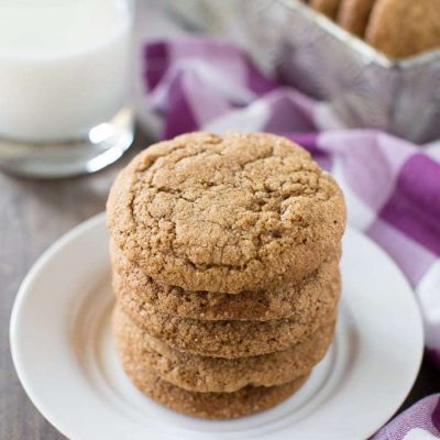 A stack of cookies sitting on a white plate with a glass of milk in the background.