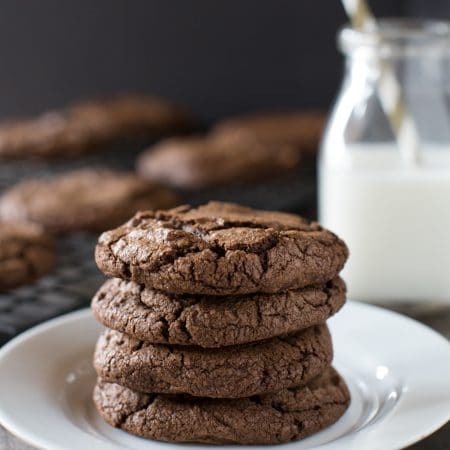 Dark chocolate cookies sitting on a white plate with a jar of milk next to them.