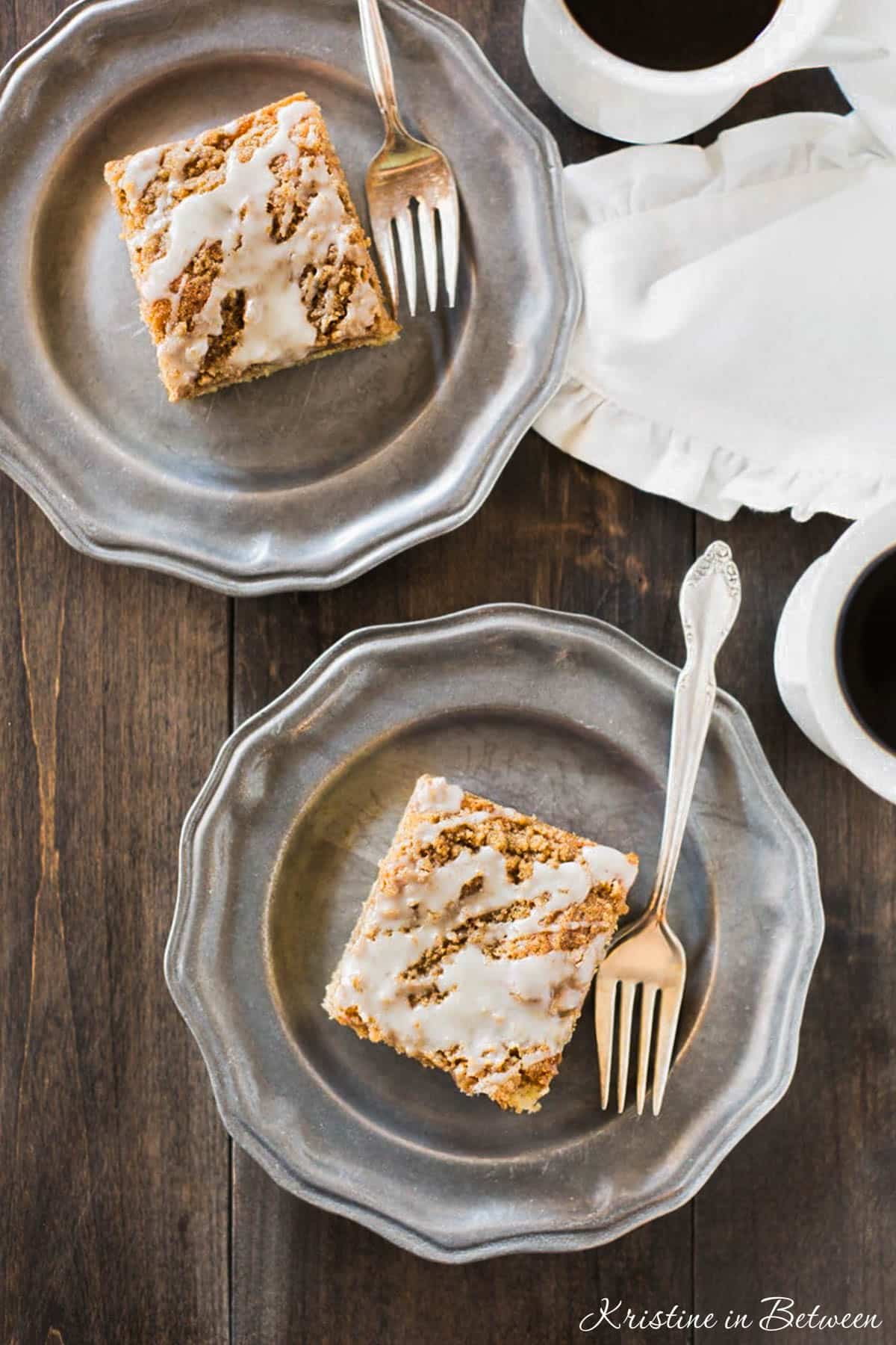 Two slices of banana crumb cake with glaze on metal plates with a cup of coffee in the background.