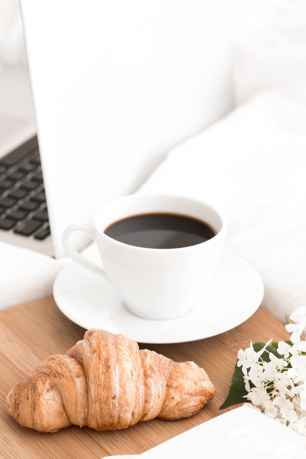A cup of coffee, a laptop, and a croissant sitting on a bed.
