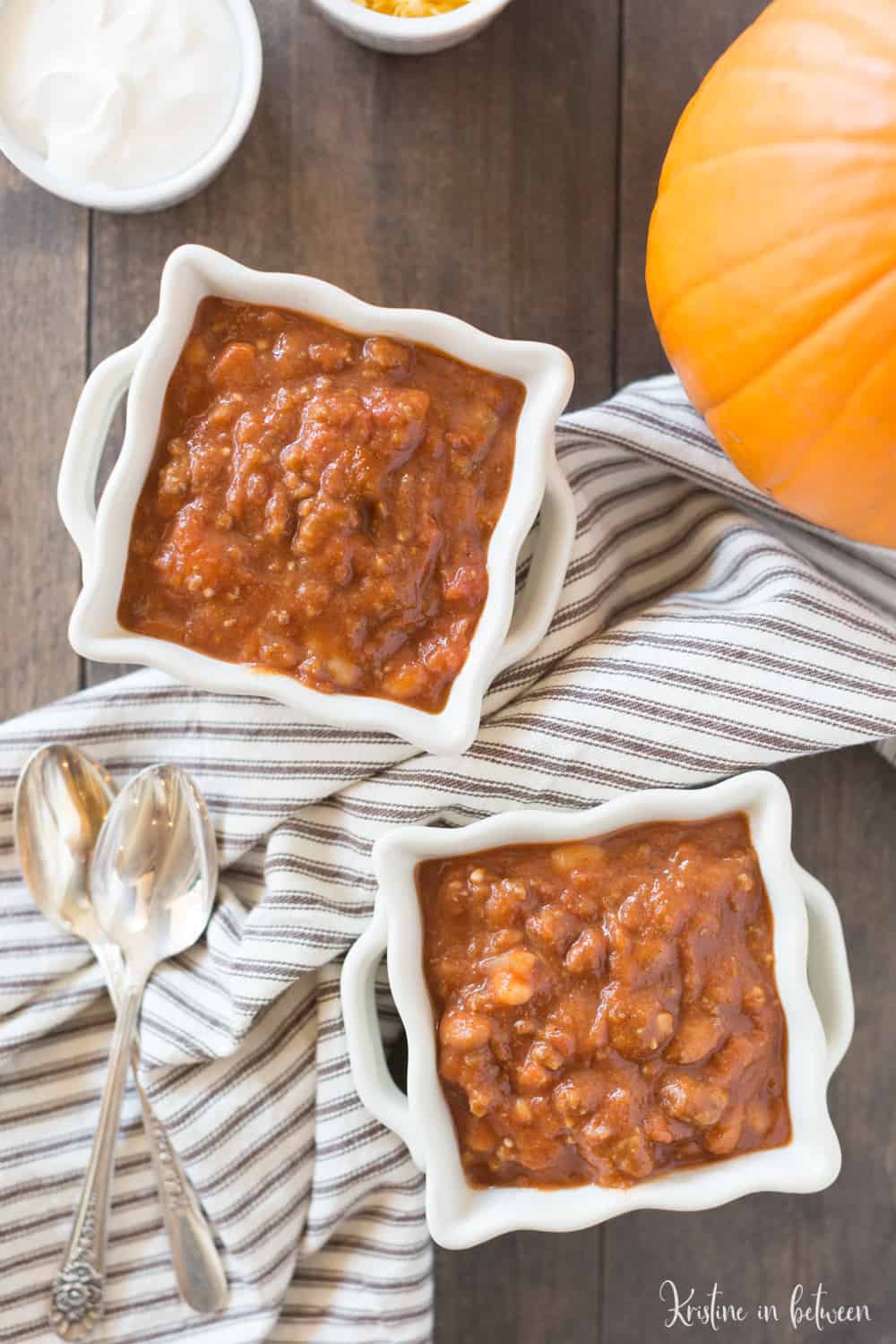 Two bowls of pumpkin chili with a small pumpkin and a brown striped napkin sitting next to them.