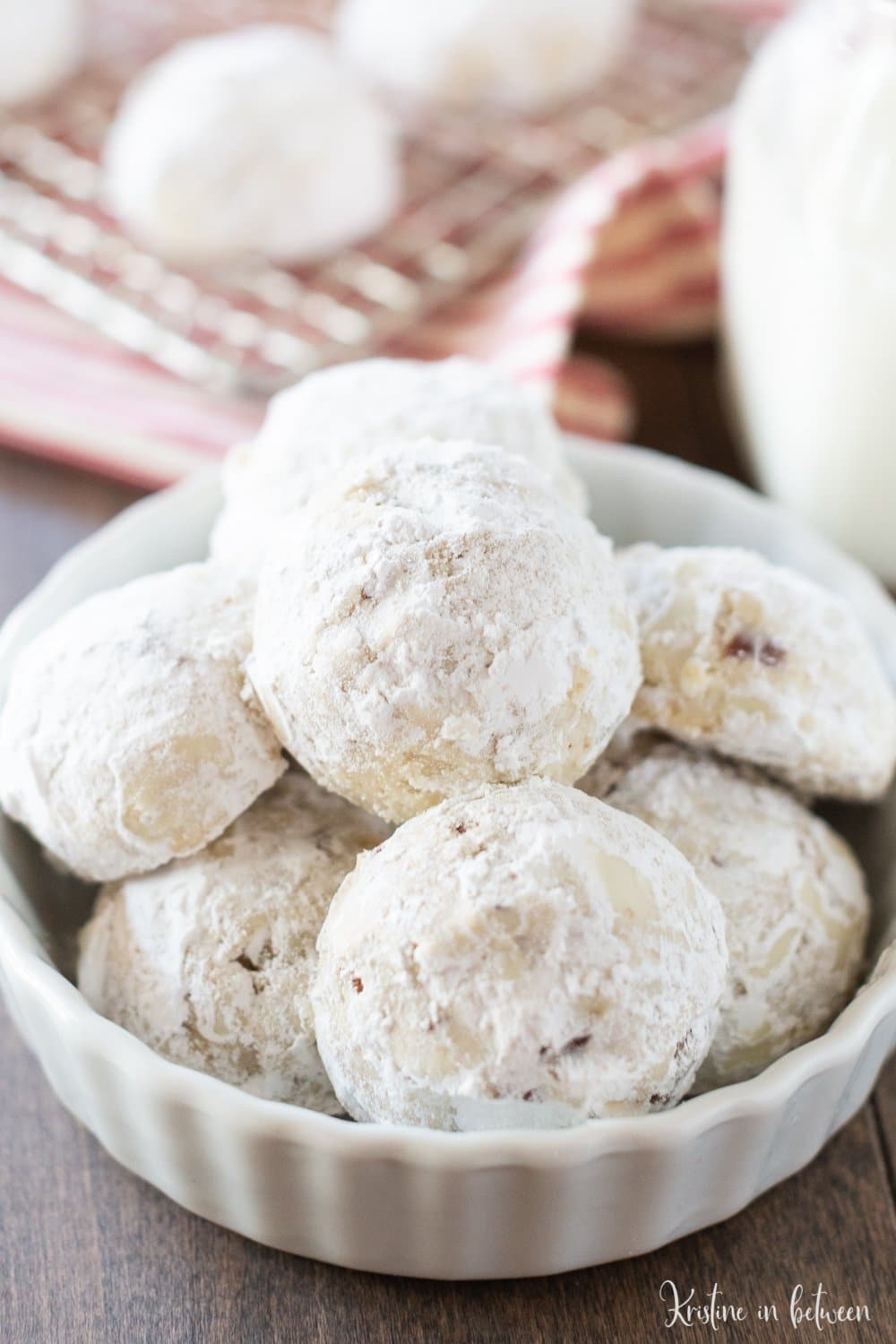 A closeup of cookies covered in powdered sugar sitting in a white bowl with a jar of milk next to them.