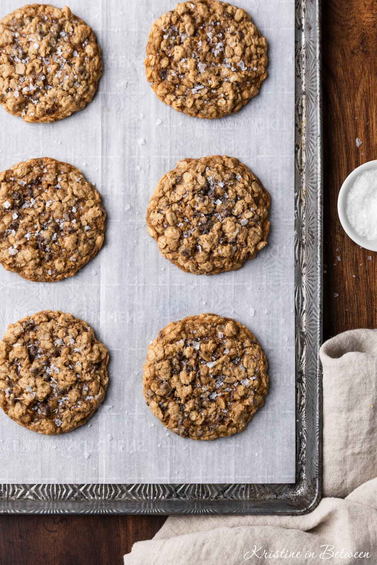 Oatmeal dark chocolate chunk cookies sitting on an antique baking tray with a small bowl of salt next to them.