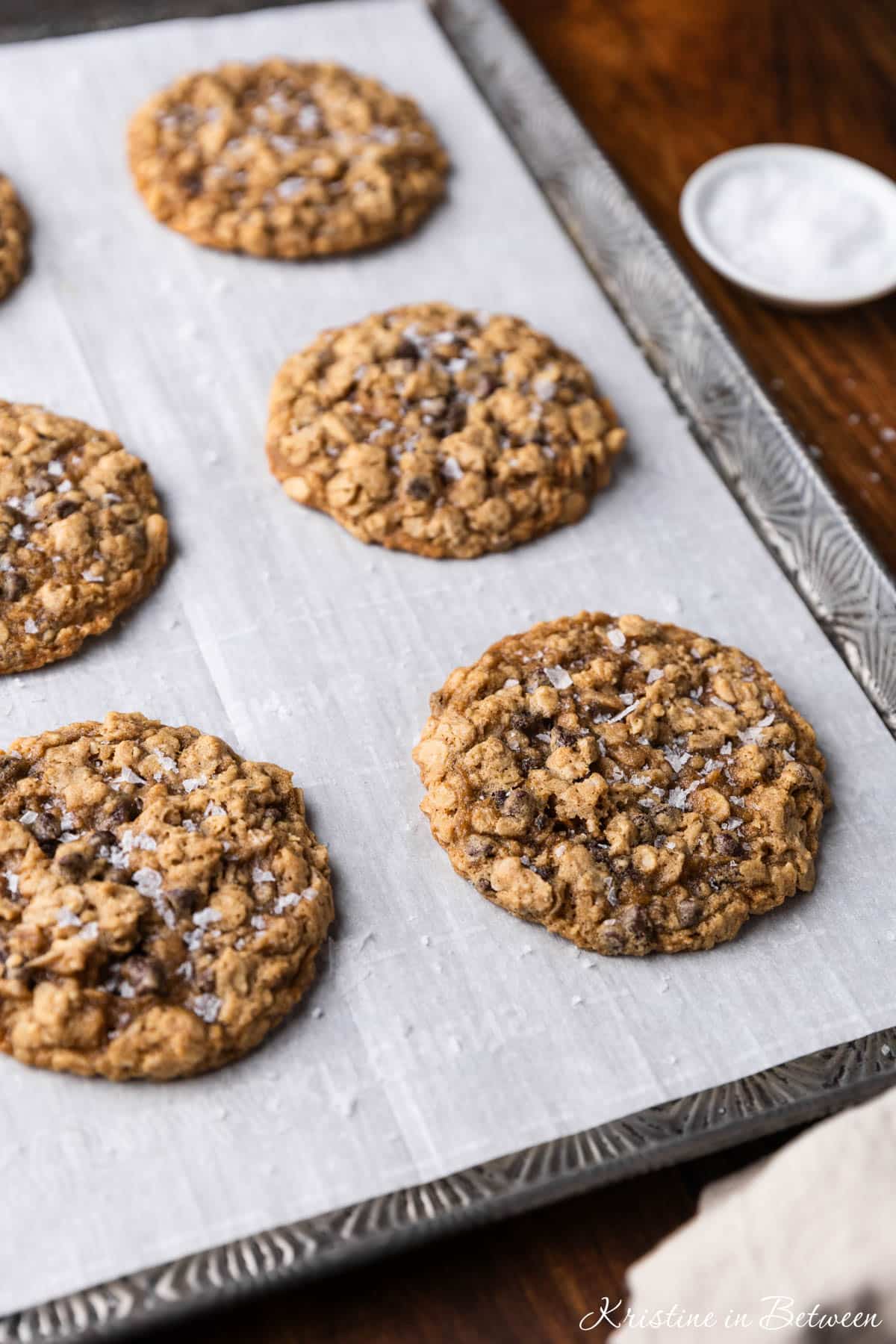 Oatmeal dark chocolate chunk cookies sitting on an antique baking tray with a small bowl of salt next to them.