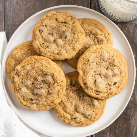 Chocolate chip cookies laying on a white plate with an eyelet napkin and a glass of milk in the background.
