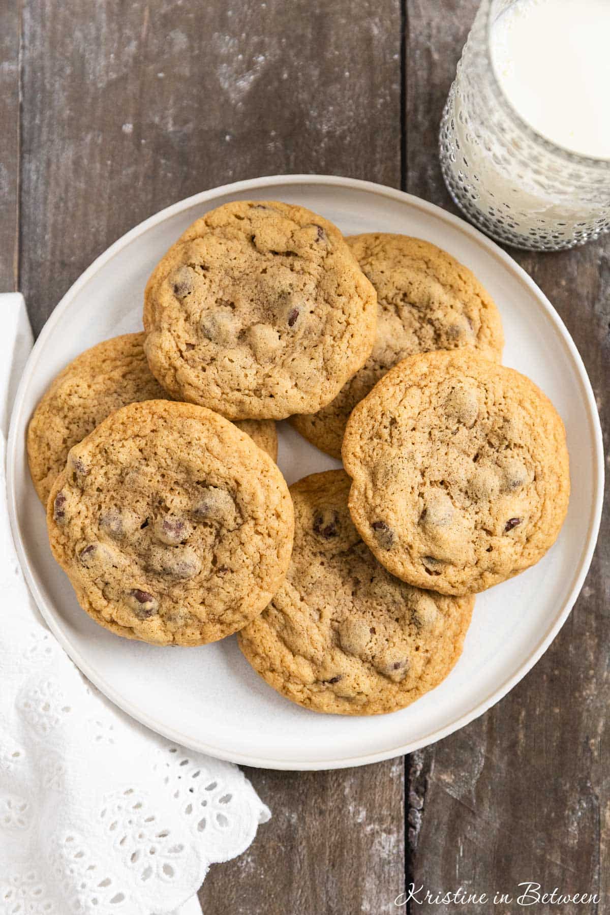 Chocolate chip cookies laying on a white plate with an eyelet napkin and a glass of milk in the background.