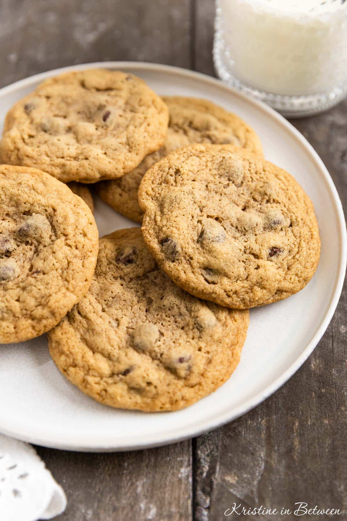 Chocolate chip cookies laying on a white plate with an eyelet napkin and a glass of milk in the background.