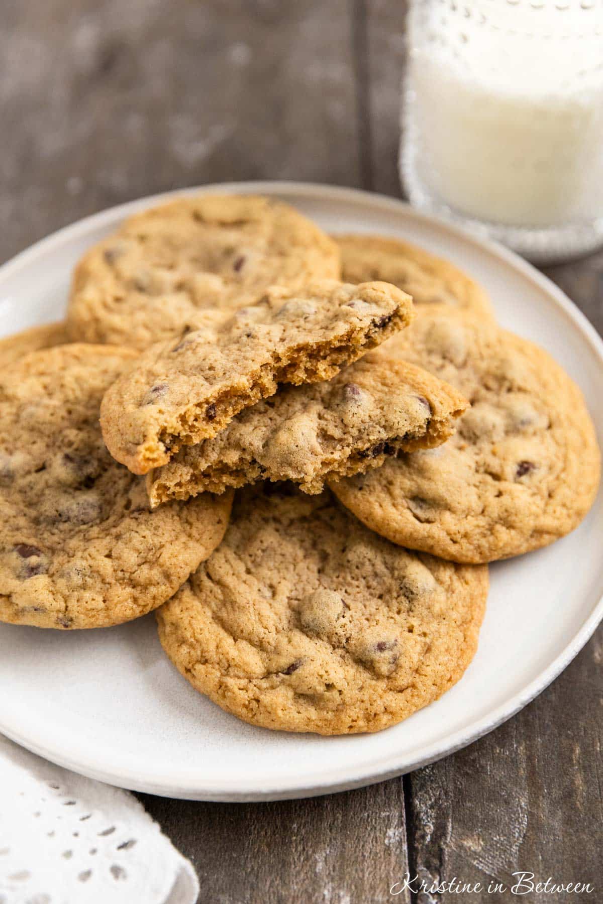Chocolate chip cookies laying on a white plate with an eyelet napkin and a glass of milk in the background.