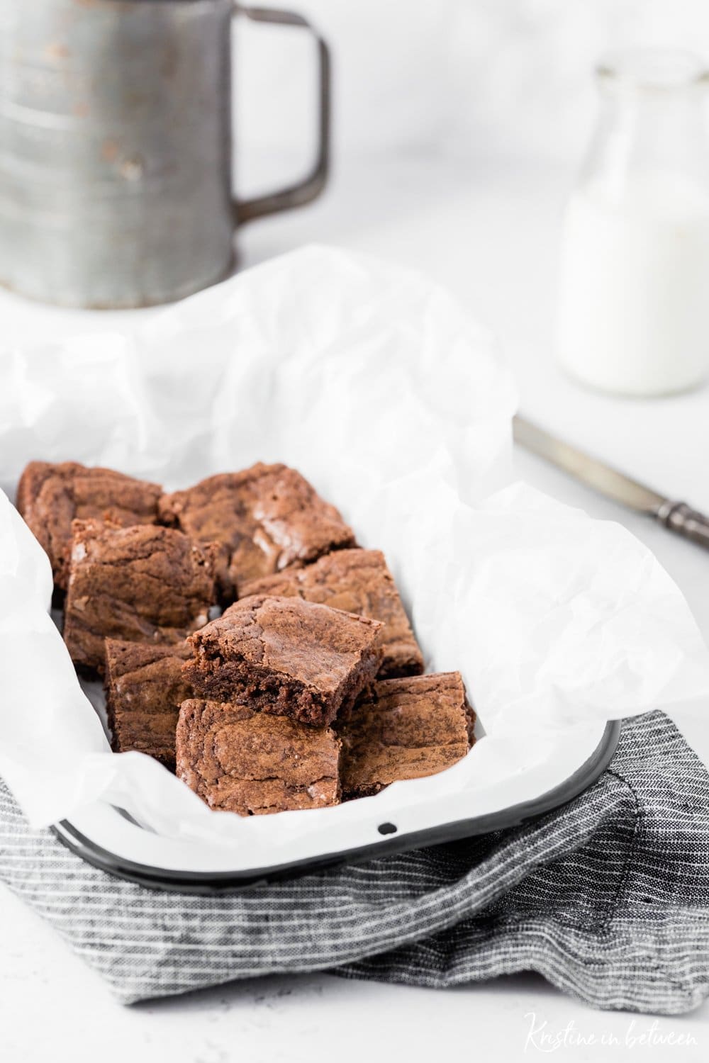 Cut up brownies laying in a white tin with an antique knife and a jar of milk in the background.