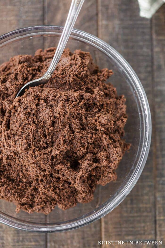 A glass bowl of chocolate cookie dough with a spoon in it.
