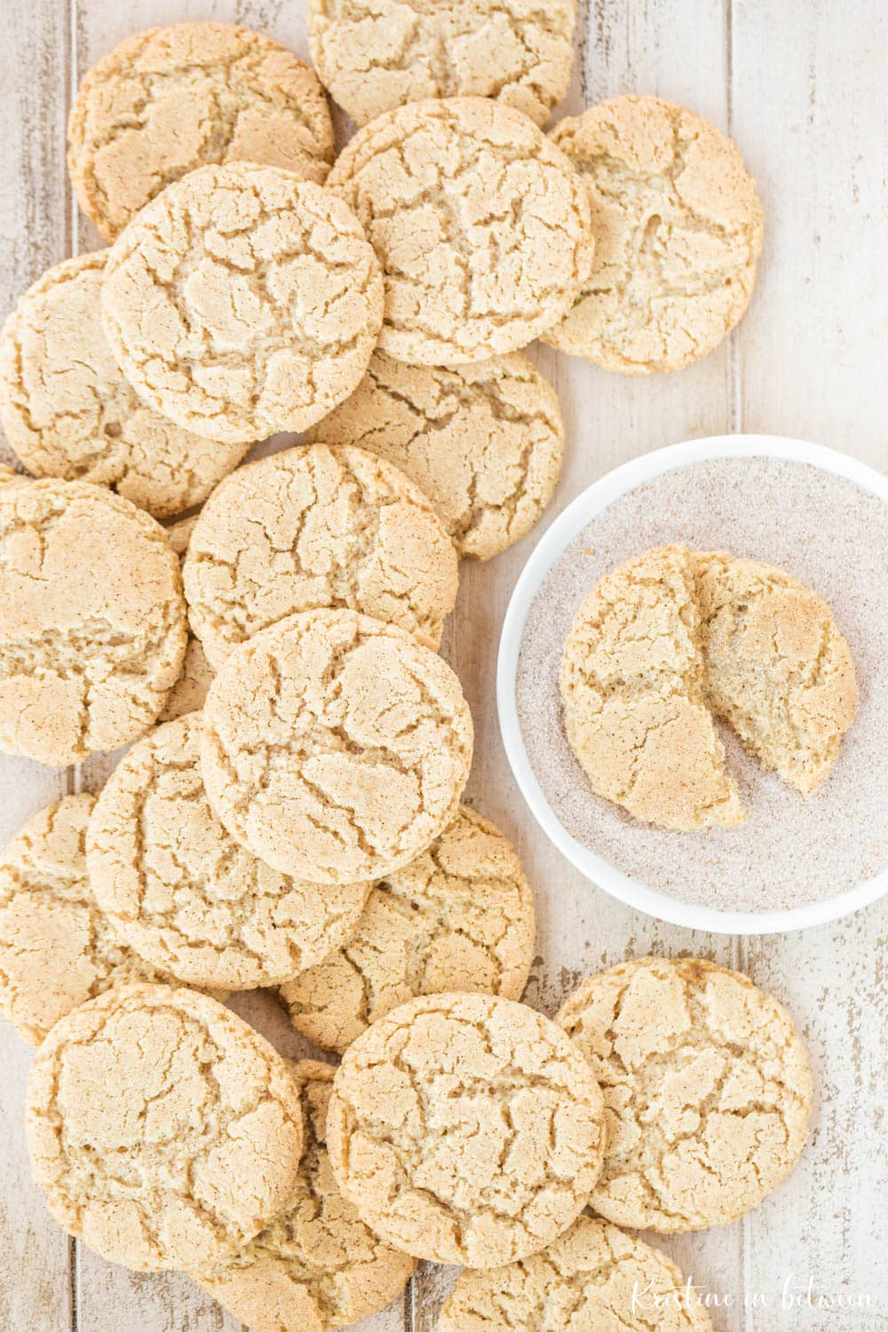Cookies piled up on a white wooden table with one sitting in a bowl of cinnamon and sugar.