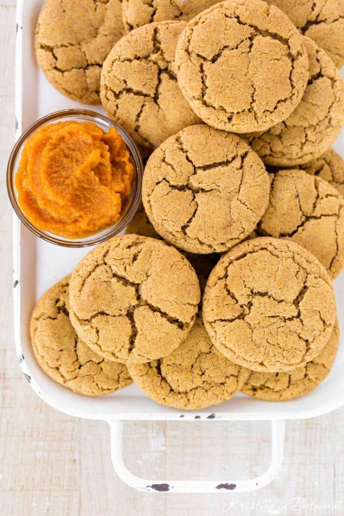 Pumpkin snickerdoodle cookies laying on a white tray with a small bowl of pumpkin puree next to them.