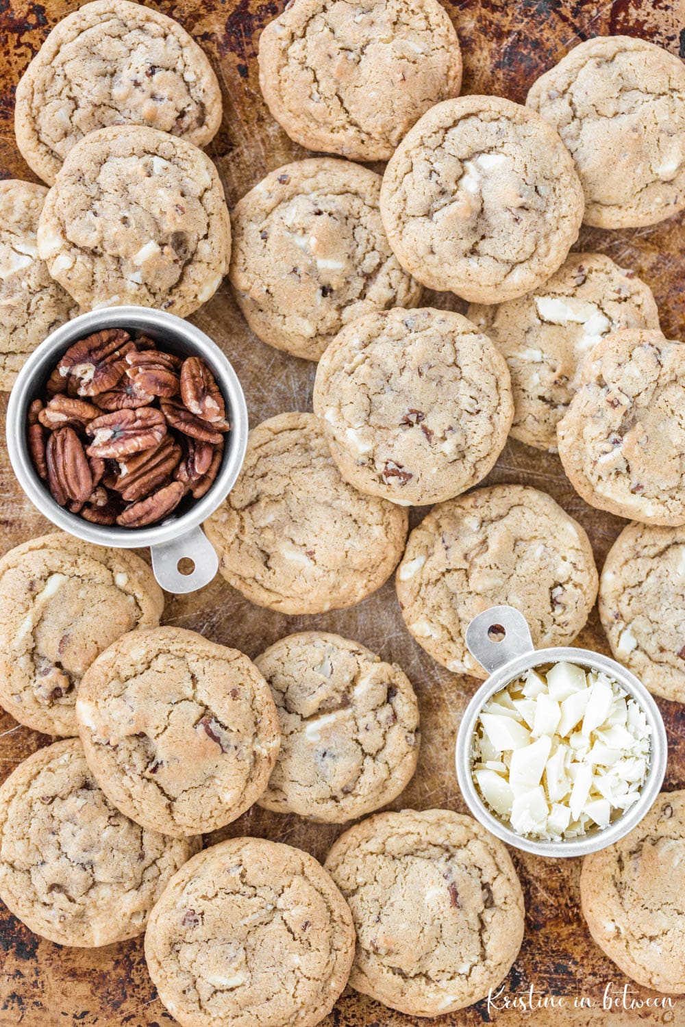 Cookies piled up on a baking sheet with a small measuring cup full of pecans and another full of chopped white chocolate.