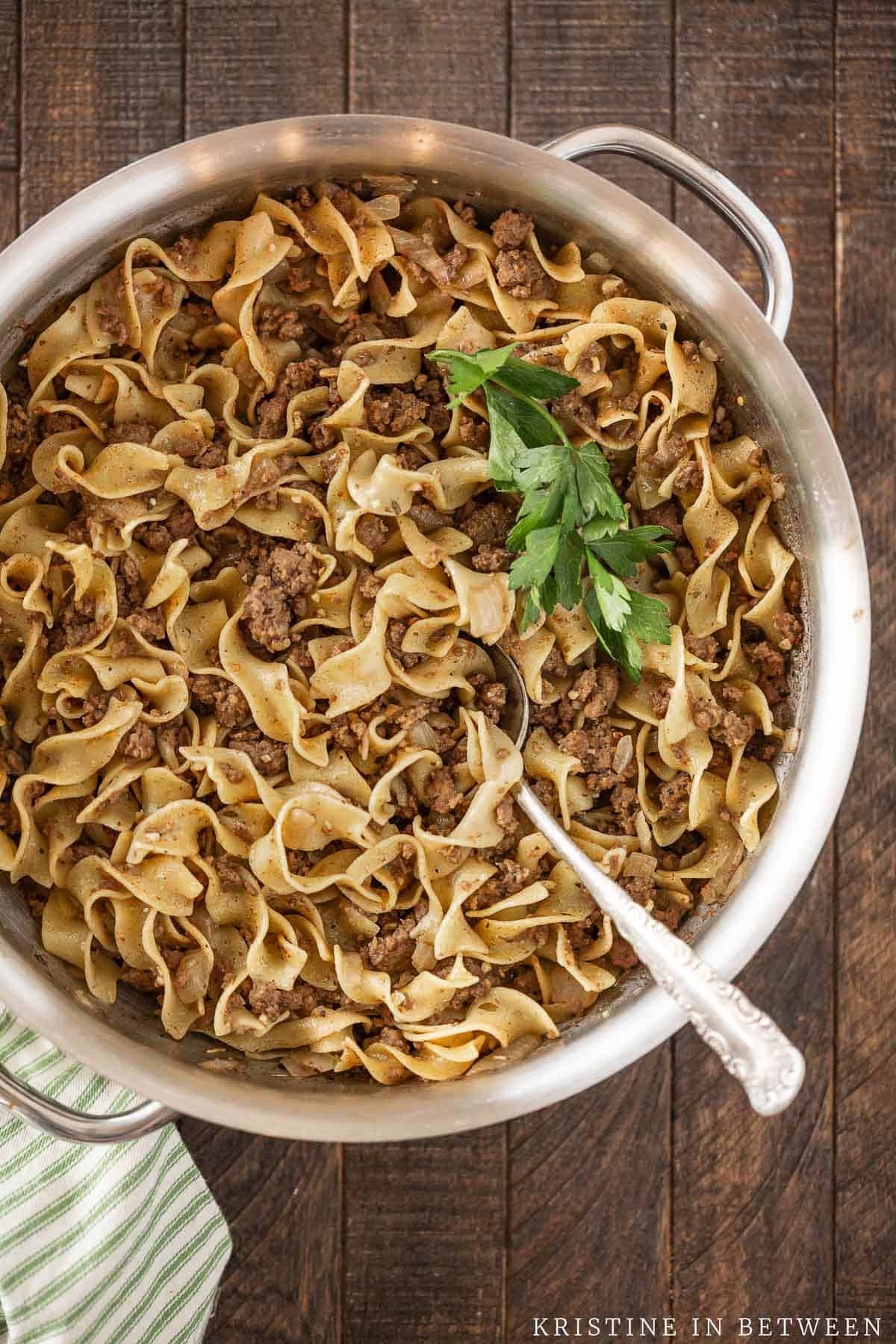 A pan of cooked beef and noodles, garnished with a fresh leaf of parsley.