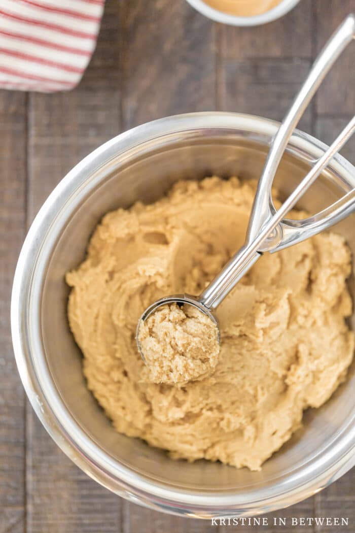 A bowl of peanut butter cookie dough with a cookie scoop in it and a small bowl of peanut butter sitting next to it.