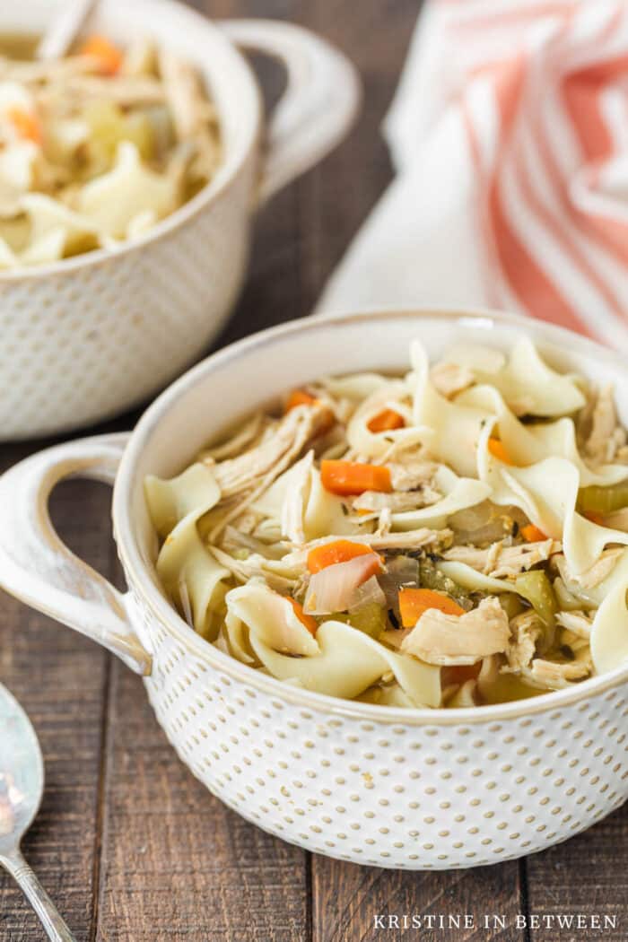 Two bowls of chicken noodle soup with antique spoons and an orange striped napkin in the background.