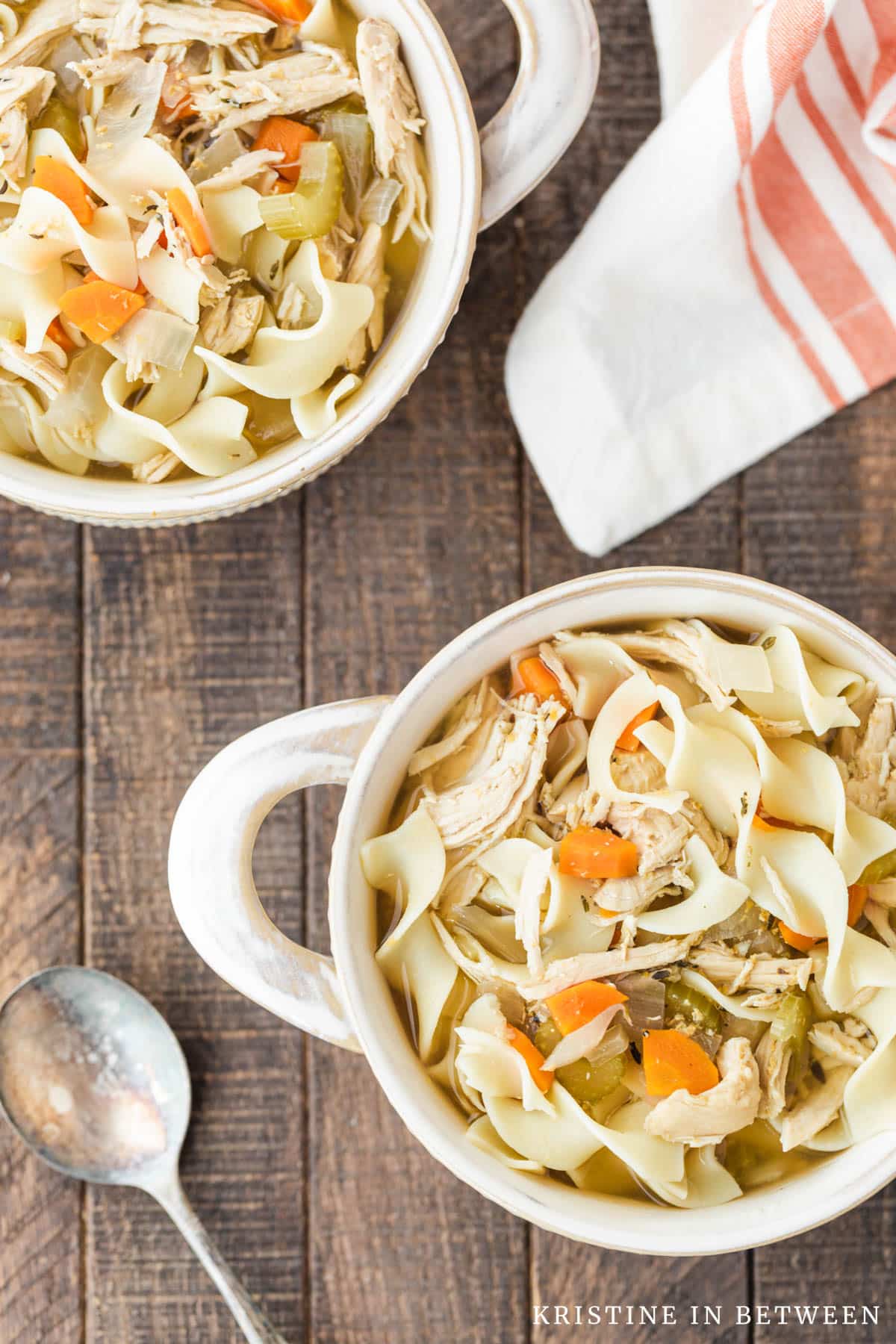 Two bowls of chicken noodle soup with antique spoons and an orange striped napkin in the background.