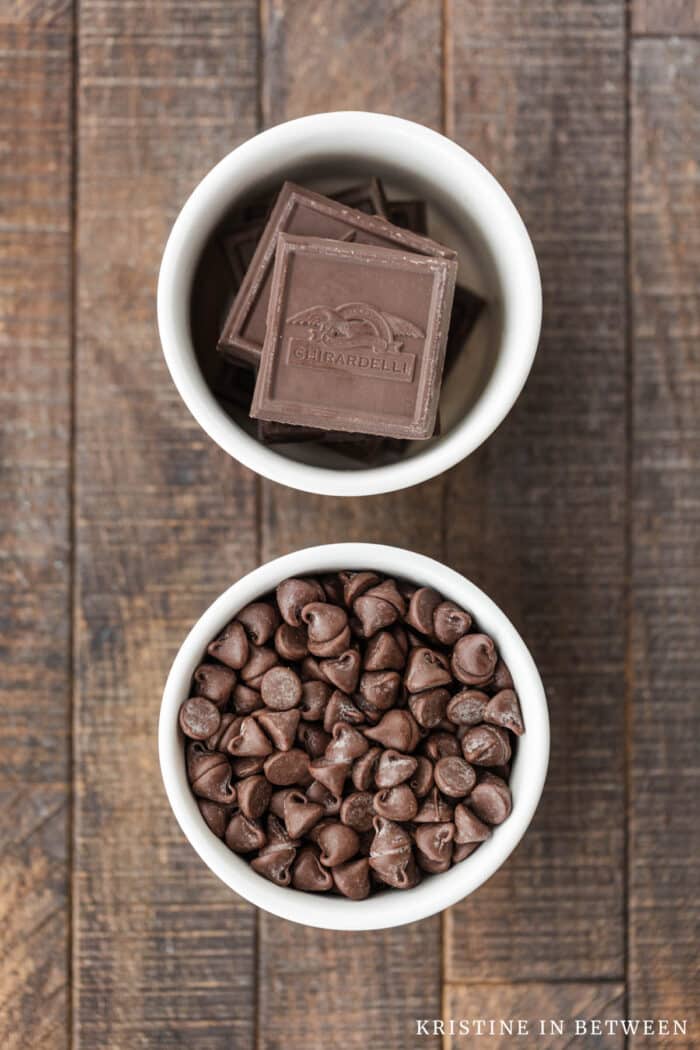 Two bowls of chocolate sitting on a wooden table, one with baking chocolate and one with chocolate chips.