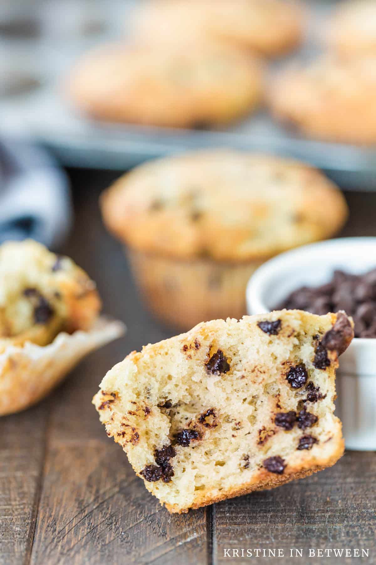 A muffin that's cut in half leaning up on a bowl of chocolate chips with more muffins in the background.