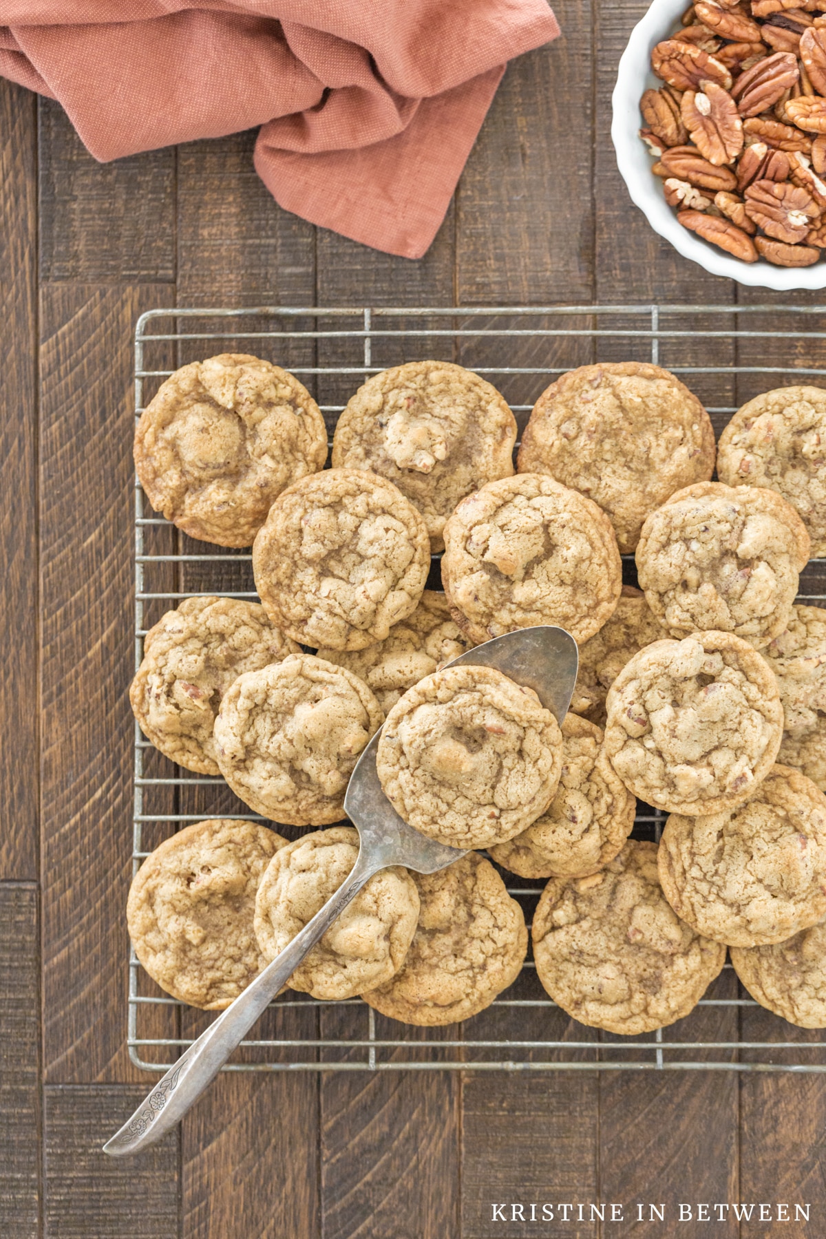 Cookies piled up on a wire rack with an antique spatula and a small bowl of pecans.