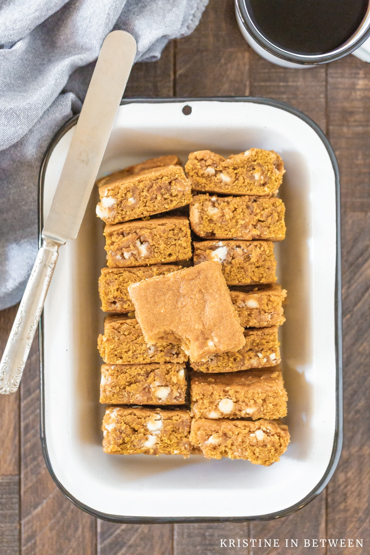 Blondies with white chocolate chips laying in a white tin pan with a knife on top of them and a cup of coffee in the background.