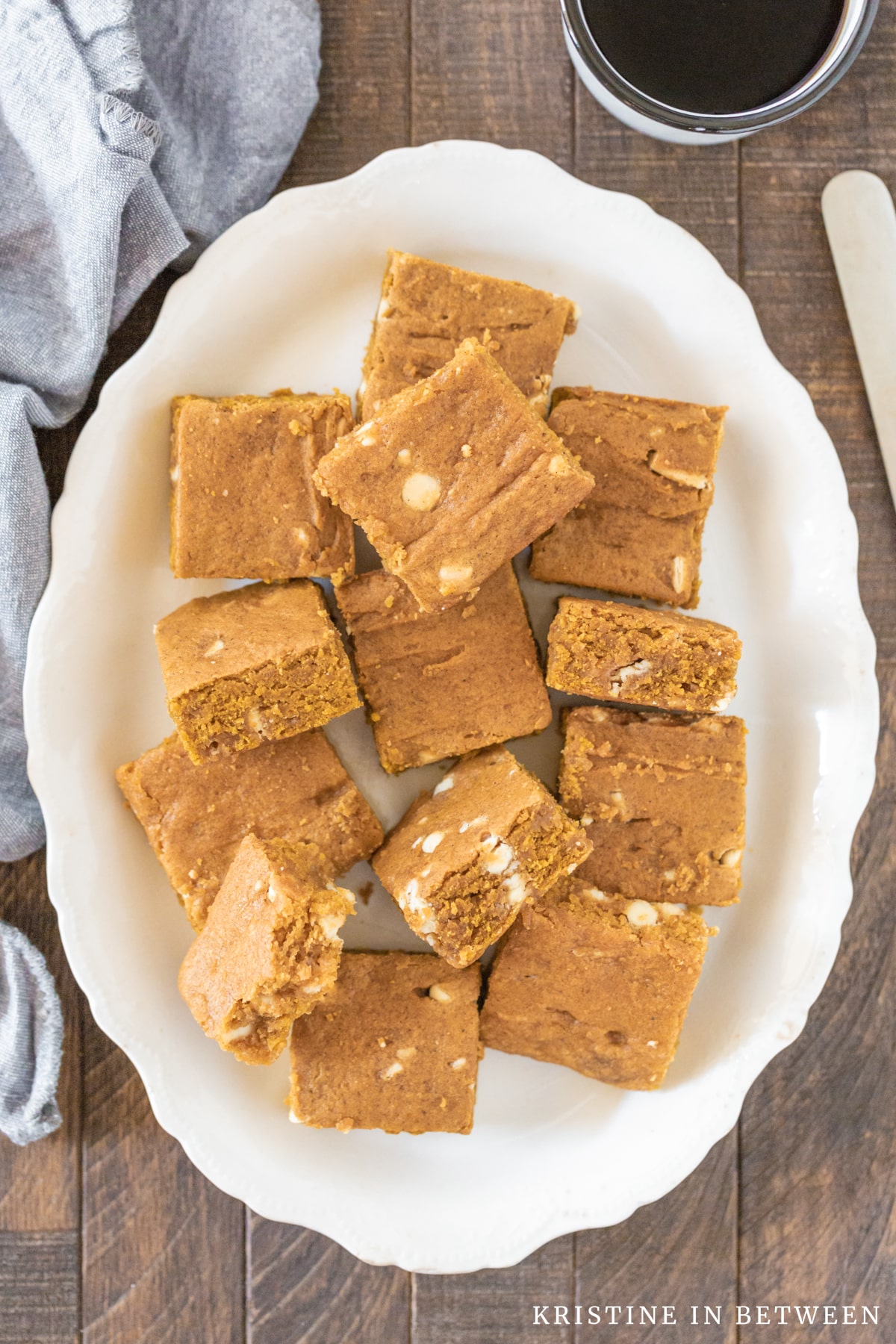 Blondies cut into squares laying randomly on a white plate with a knife next to them.