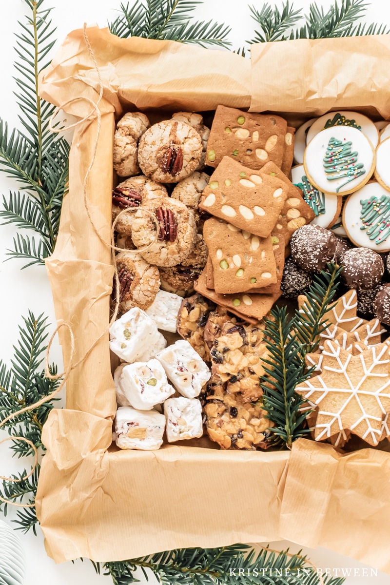 A variety of Christmas cookies packed up in a box with some fern branches and brown twin next to them.