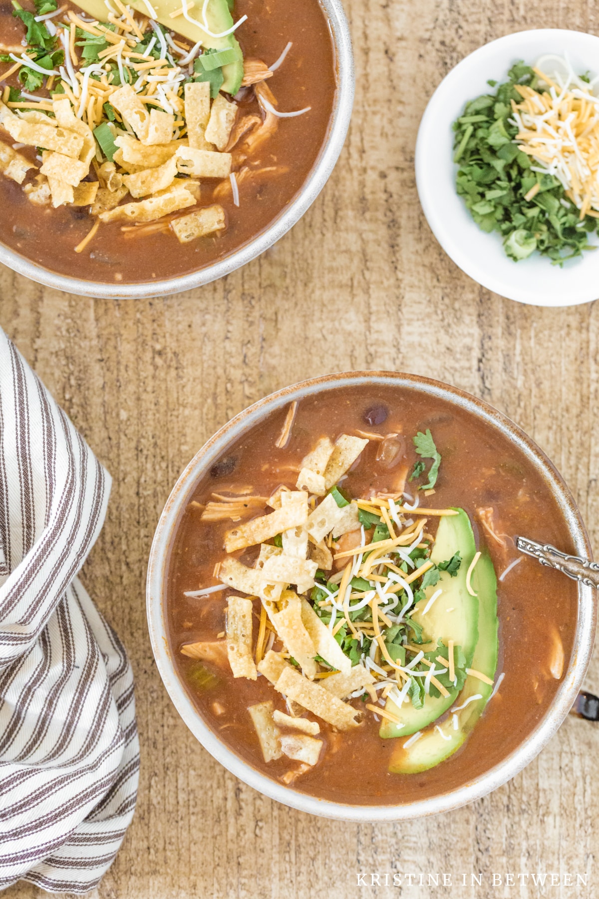 Two bowls of enchilada soup topped with cheese, tortilla strips, and avocado sitting with a striped napkin.