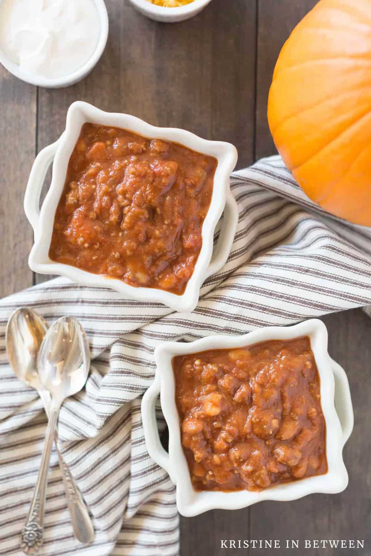 Two bowls of pumpkin chili with a small pumpkin and a brown striped napkin sitting next to them.