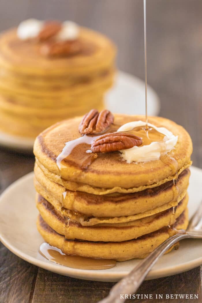 Pumpkin spice pancakes stacked upon a white plate with syrup, pecans, and butter on top.