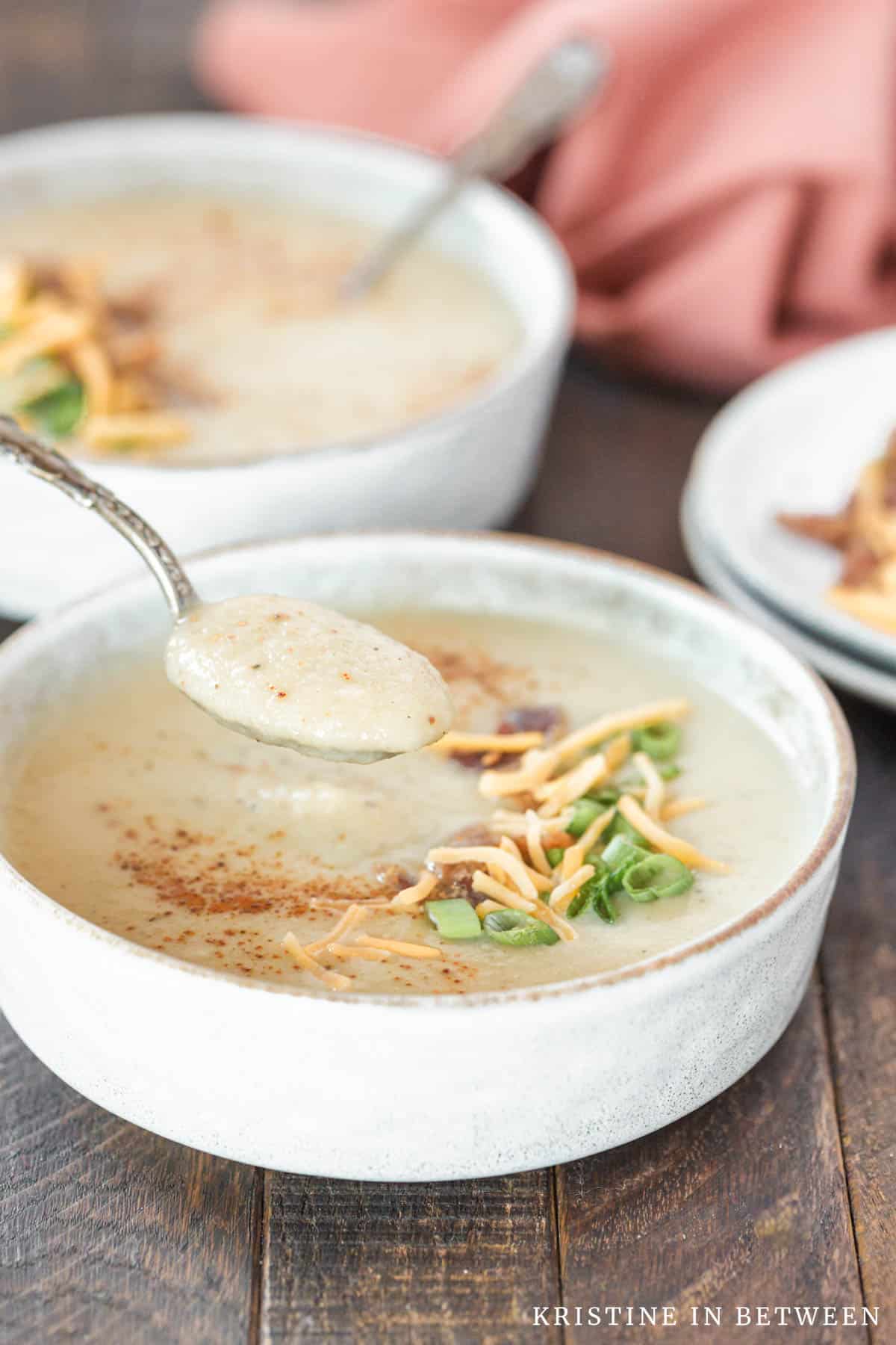 A bite of cauliflower soup on a spoon with a second bowl of soup in the background.
