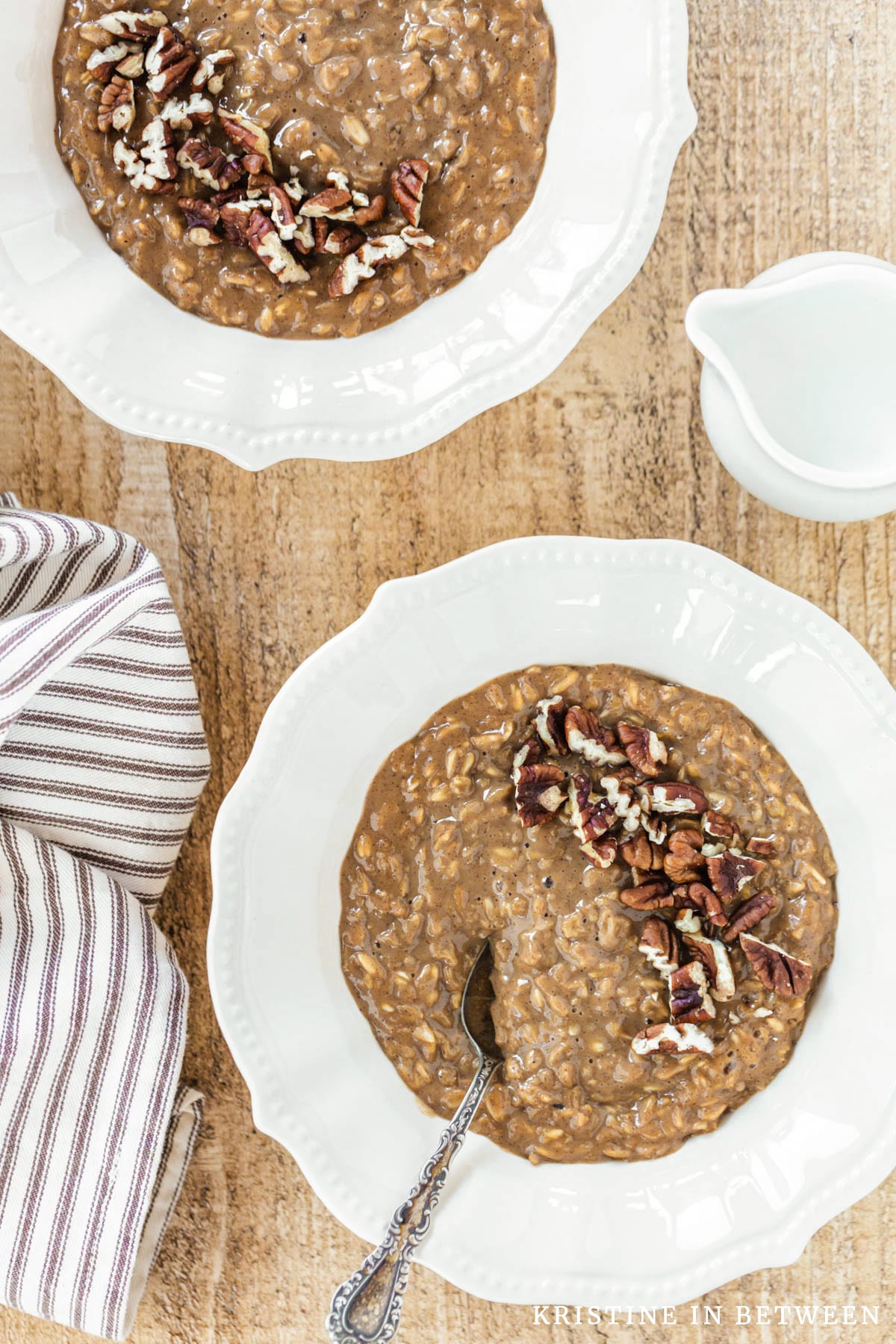 A bowl of gingerbread oatmeal topped with chopped pecans sitting with a spoon and a small pitcher of milk.