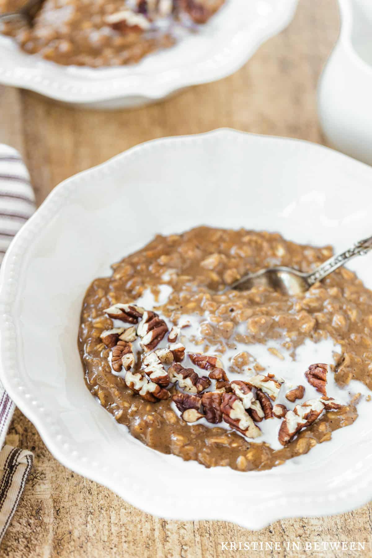 A bowl of gingerbread oatmeal topped with chopped pecans sitting with a spoon and a small pitcher of milk.