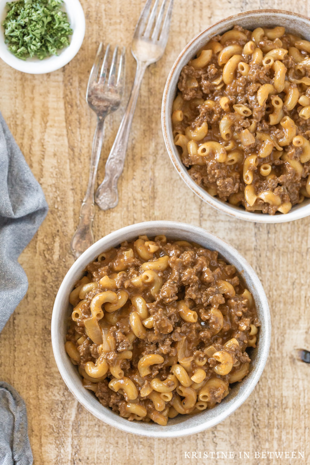 Two bowls of chili mack sitting with two forks and a blue napkin.