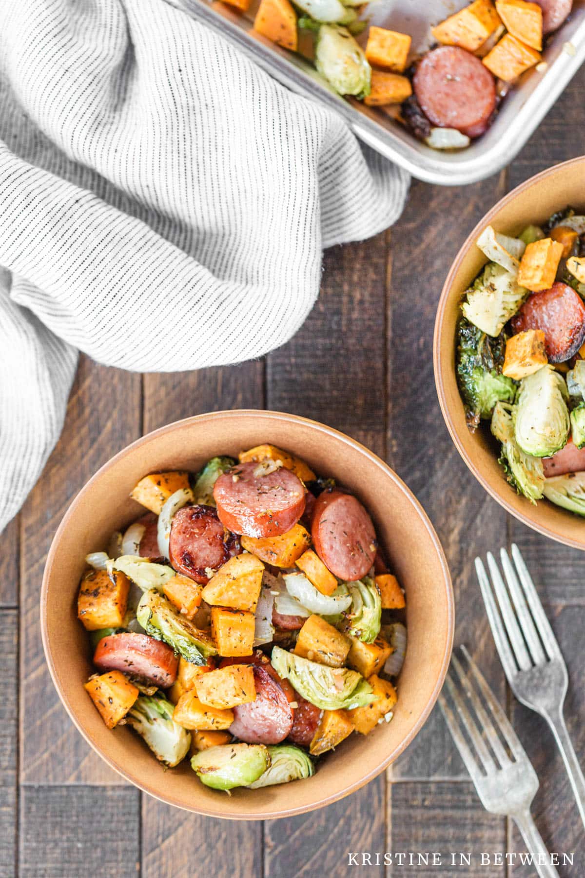 A bowl of sheet pan Kielbasa sausage and Brussel sprouts with two forks and a sheet pan in the background.