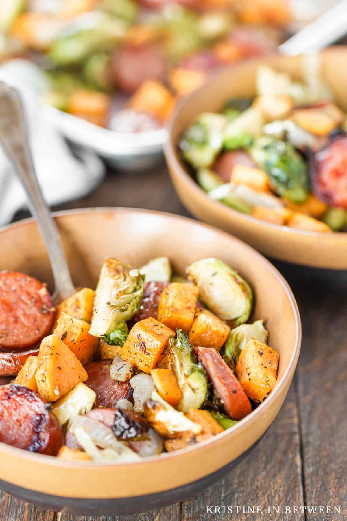 Two bowls of kielbasa and Brussel sprouts in a bowl with a sheet pan in the background.