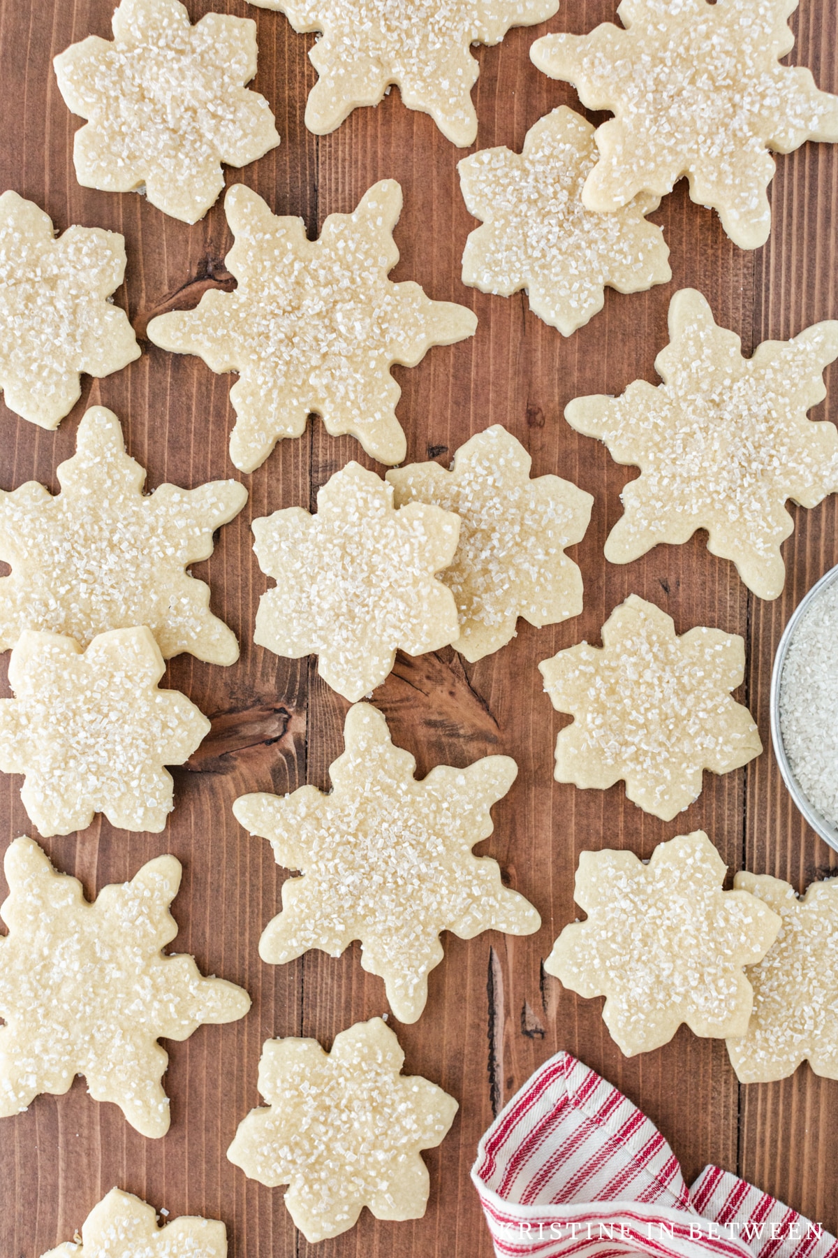 Cut out sugar cookies laying on a wooden table with a small bowl of sugar.