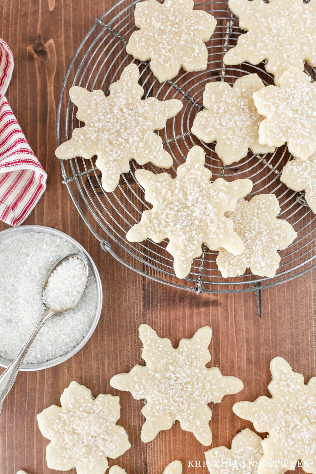 Cut out sugar cookies laying on a wooden table with a few on a circle rack and a bowl of sugar next to them.