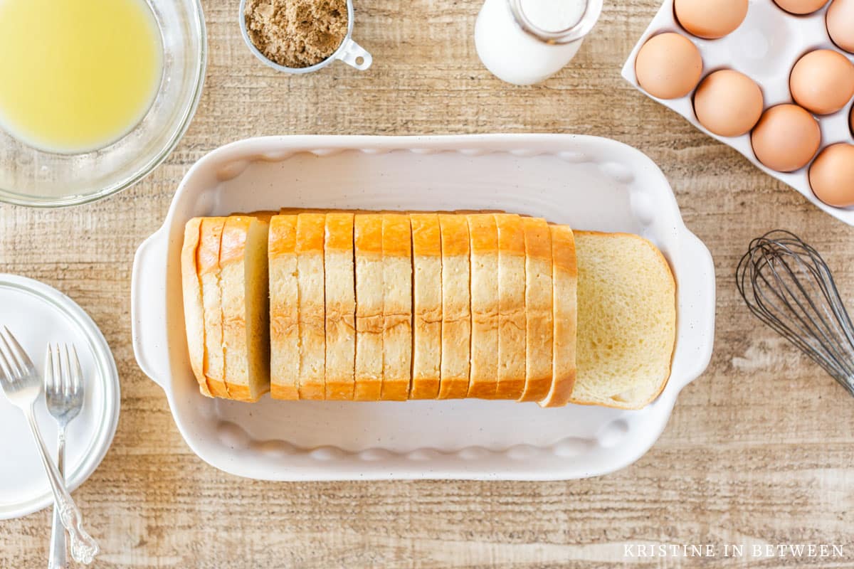 A loaf of brioche bread in a casserole dish with eggs, milk, and a whisk sitting next to it.