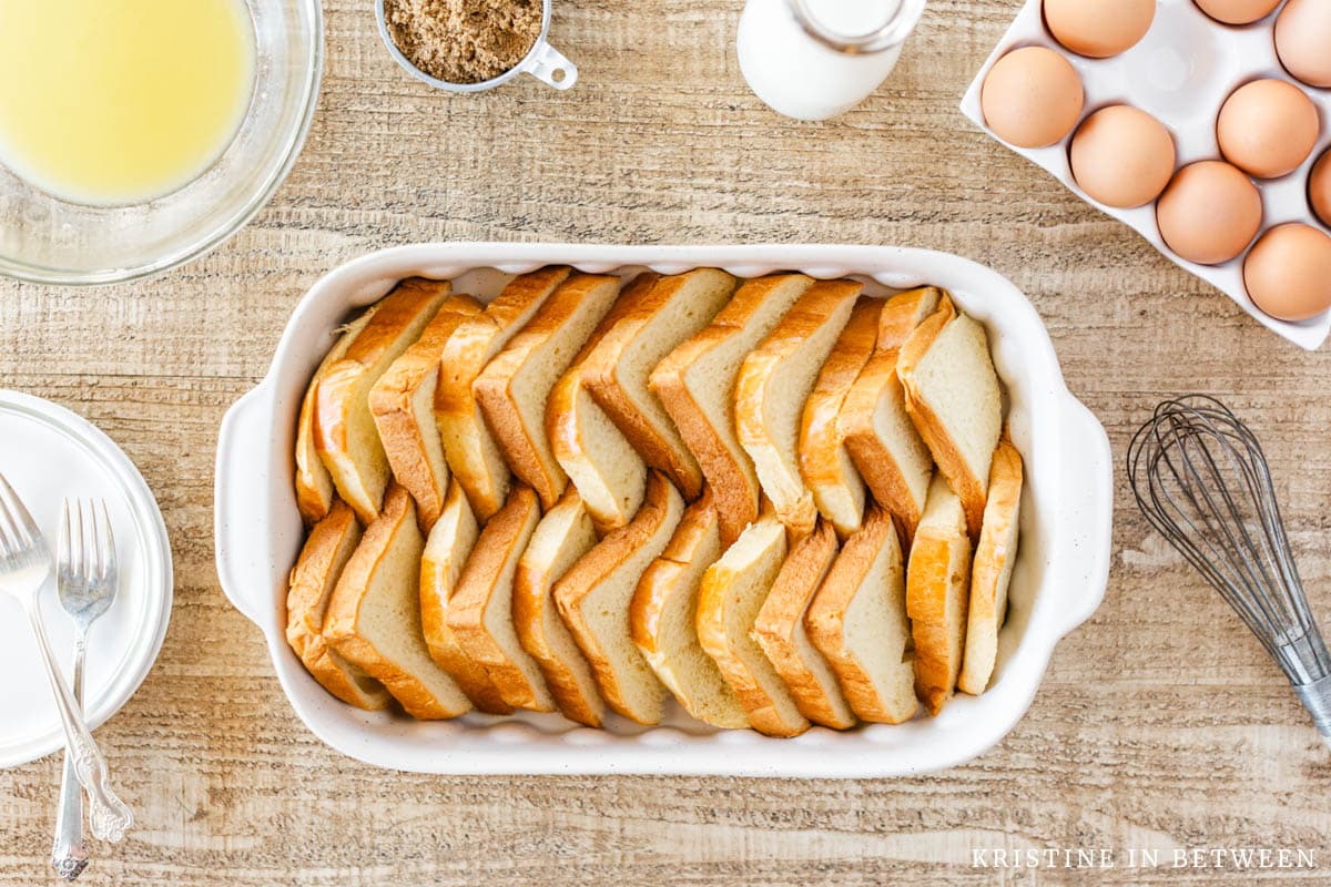 Triangles of bred arranged in a casserole dish with eggs, butter, milk and a whisk sitting next to it.