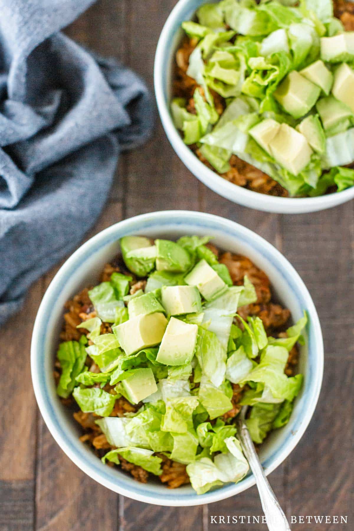 Beef burrito casserole in a white bowl, topped with chopped lettuce and avocado with a blue napkin sitting next to it.