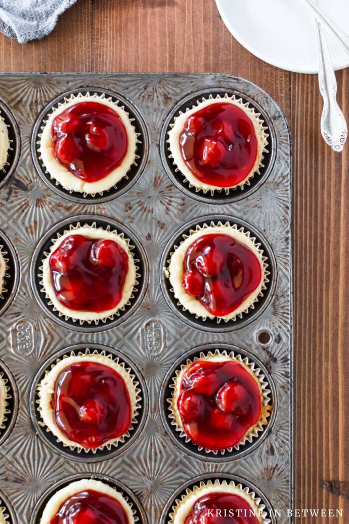 A muffin tin full of cherry tarts with plates and forks in the background.