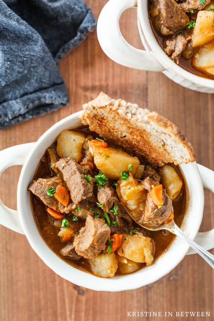 A bowl of beef stew with a spoon sitting on top and a slice of bread in the bowl.