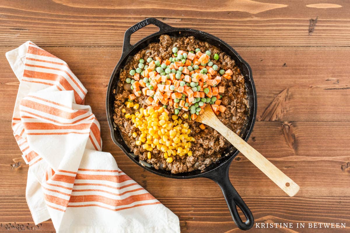 Cooked beef in a cast iron skillet with peas, corn, and carrots on top.