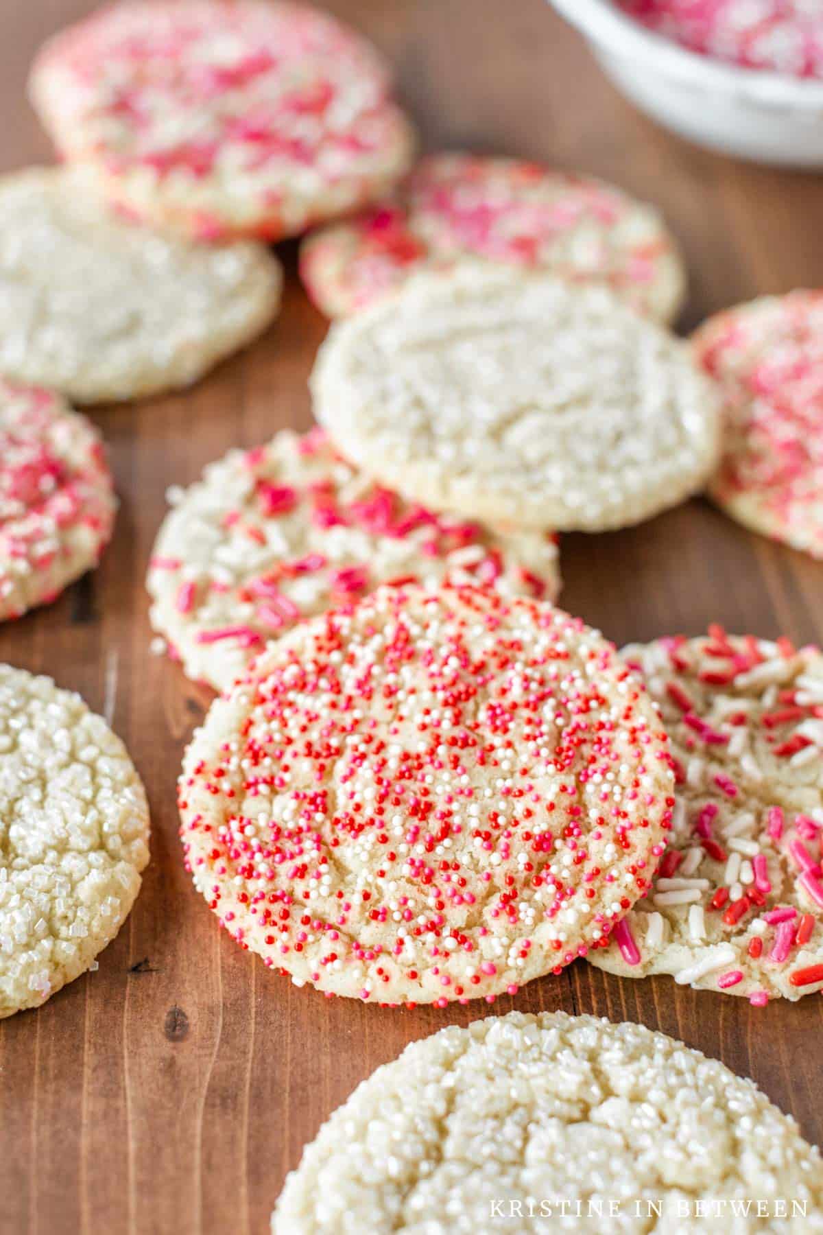Valentine sugar cookies with red, white, and pink sprinkles laying on a wooden board with a small bowl of sprinkles.