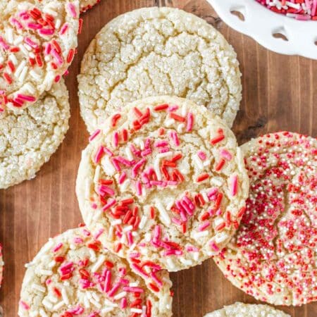 Valentine sugar cookies with red, white, and pink sprinkles laying on a wooden board with a small bowl of sprinkles.