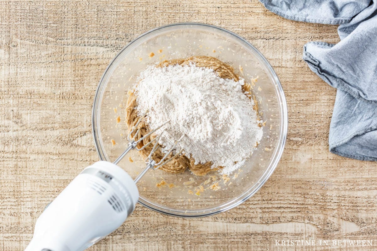 Flour on top of wet ingredients in a glass bowl with a white hand mixer.
