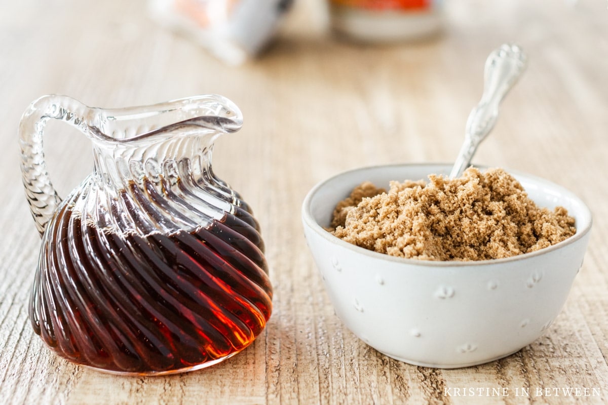 A small bowl of brown sugar with a spoon in it sitting with a jar of maple syrup.